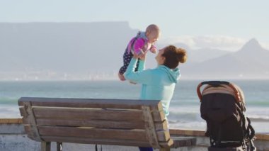 Genç anne kollarını tutuyor ve tatil sırasında gün batımında deniz kenarında bir kumsalda yeni doğmuş bir bebekle oynuyor..