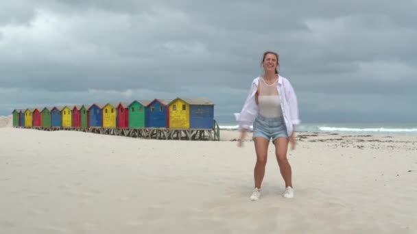 Девушка танцует на белых песках Южной Африки рядом с красочным лесом Muizenberg Beach Houses. — стоковое видео