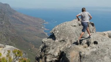 Camera follows boy running up on top of mountain summit, jumps on top of rocks, raises arms into air, happy and drunk on life, travel and happiness.