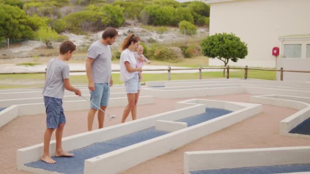 Famille de quatre personnes jouant sur un terrain de golf miniature, garçon faisant du putt dans un trou. — Video