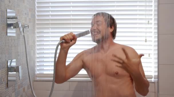 Ein junger gutaussehender Mann mit dunklen Haaren duscht und singt emotional wie in ein Mikrofon. Wasserstrahlen fließen über Gesicht und Brust. — Stockvideo