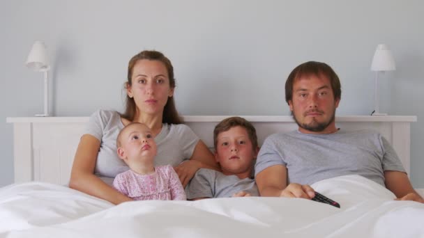 Šťastná mladá evropská rodina se dvěma dětmi se shromáždí na gauči a dívají se na televizi v badroomu s úsměvem. Togetherness a zábava. — Stock video