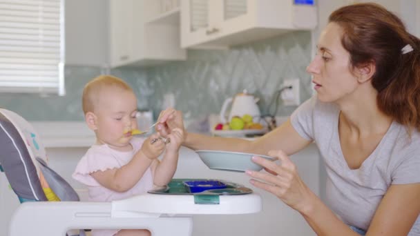 Precioso niño en la silla de bebé en casa está disfrutando de su comida sólida de cuchara servida por su madre a la hora del almuerzo. — Vídeos de Stock