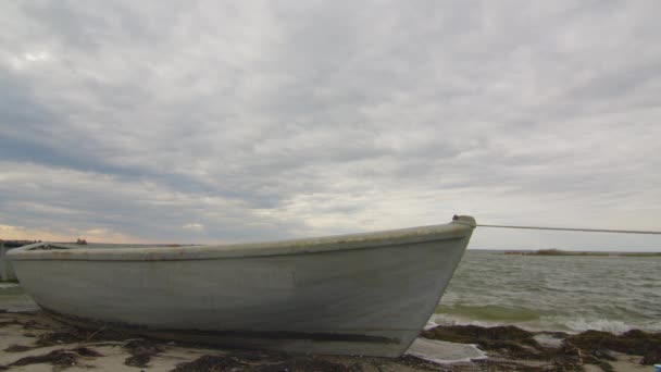 A wooden fishing boat moored on a sandy and grassy shore at sunset with a cloudy sky, time lapse. — Vídeos de Stock