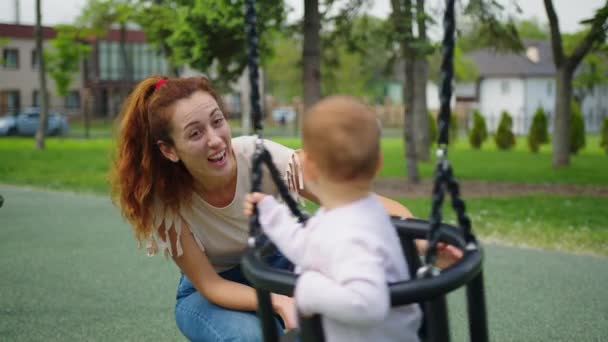 Lycklig familj mamma rullar det lilla barnet i flykt på swing i parken. På helgerna går mor och dotter. Familjepromenader i parken. Ung och ung förälder. begreppet barn och familj. Lagarbete. — Stockvideo