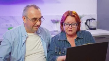 Mutlu orta yaşlı çift dizüstü bilgisayarlarını kullanarak alışveriş yapıyor, yetişkin aile içi eğitimleri alıyor internet haberlerini tartışıyor, internet sitesinden seyahat teklifini seçiyor..
