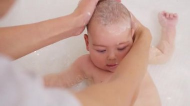 An adorable baby taking a bath with mother washing his head.