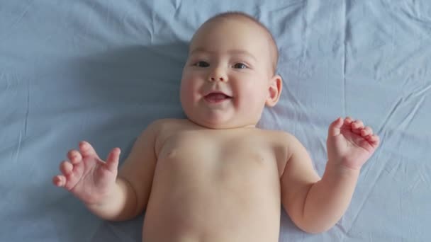 Ένα μικρό καυκάσιο νεογέννητο μωρό είναι αστείο χαμογελαστό, ξαπλωμένο πίσω στην κούνια του παιδιού. Πορτρέτο ενός παιχνιδιάρικου και δραστήριου παιδιού σε κοντινό πλάνο. — Αρχείο Βίντεο