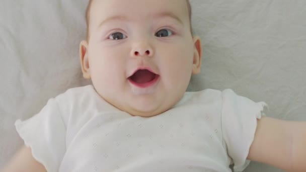 Retrato de cerca de una niña. La cara de una niña caucásica acostada en la cama. bebé 6 mounth mirando en cámara. — Vídeos de Stock