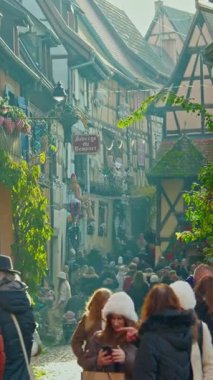Eguisheim, Fransa - 3.12.2025: Noel için dekore edilmiş küçük, güzel bir köy. İnsanlar, Fransız köyü Eguisheim 'ın güzel dekore edilmiş sokaklarında geziniyor. Dikey video.