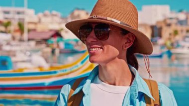 Şapkalı ve sırt çantalı bir kız eski renkli bir balıkçı kasabası olan Marsalokk 'ta ünlü çok renkli tekneleriyle yürüyor..