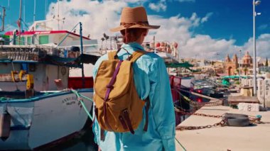 Şapkalı ve sırt çantalı bir kız eski renkli bir balıkçı kasabası olan Marsalokk 'ta ünlü çok renkli tekneleriyle yürüyor..