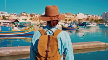 Şapkalı ve sırt çantalı bir kız eski renkli bir balıkçı kasabası olan Marsalokk 'ta ünlü çok renkli tekneleriyle yürüyor..