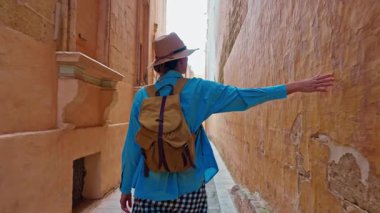 Şapkalı ve sırt çantalı bir kadın turist Mdina, Malta 'daki geleneksel dar sokaklar boyunca yürüyor..