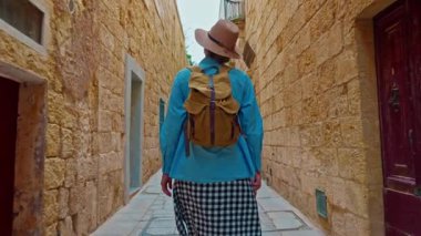 Şapkalı ve sırt çantalı bir kadın turist Mdina, Malta 'daki geleneksel dar sokaklar boyunca yürüyor..