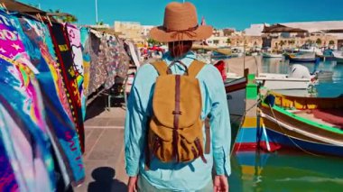 Şapkalı ve sırt çantalı bir kız eski renkli bir balıkçı kasabası olan Marsalokk 'ta ünlü çok renkli tekneleriyle yürüyor..