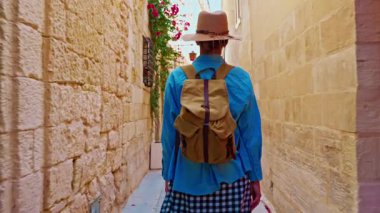 Şapkalı ve sırt çantalı bir kadın turist Mdina, Malta 'daki geleneksel dar sokaklar boyunca yürüyor..