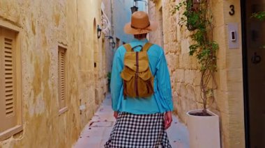 Şapkalı ve sırt çantalı bir kadın turist Mdina, Malta 'daki geleneksel dar sokaklar boyunca yürüyor..