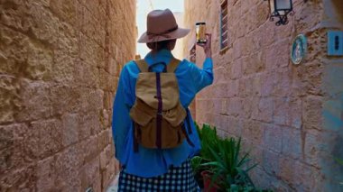 Şapkalı ve sırt çantalı bir kadın turist Mdina, Malta 'daki geleneksel dar sokaklar boyunca yürüyor..