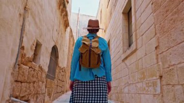 Şapkalı ve sırt çantalı bir kadın turist Mdina, Malta 'daki geleneksel dar sokaklar boyunca yürüyor..