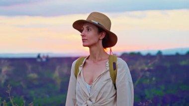 A girl tourist with a hat and a backpack walks through a lavender field during a beautiful sunset in Provence, France.