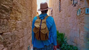 Şapkalı ve sırt çantalı bir kadın turist Mdina, Malta 'daki geleneksel dar sokaklar boyunca yürüyor..