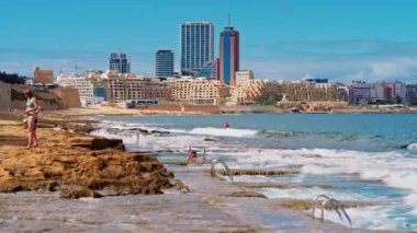 Sliema, Malta: Rocky plajları ve Sliema 'nın gezinti güvertesi. İnsanlar şehrin arka planında kayalık sahilde yüzüyor ve güneşleniyor..