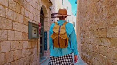 Şapkalı ve sırt çantalı bir kadın turist Mdina, Malta 'daki geleneksel dar sokaklar boyunca yürüyor..