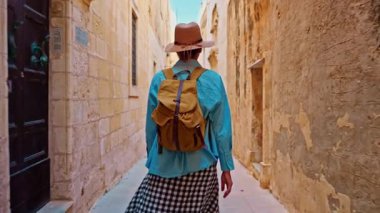 Şapkalı ve sırt çantalı bir kadın turist Mdina, Malta 'daki geleneksel dar sokaklar boyunca yürüyor..