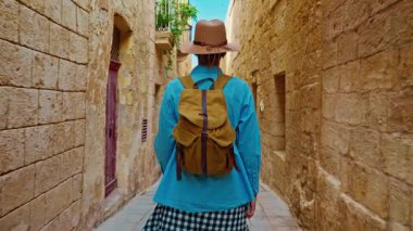 Şapkalı ve sırt çantalı bir kadın turist Mdina, Malta 'daki geleneksel dar sokaklar boyunca yürüyor..