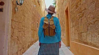 Şapkalı ve sırt çantalı bir kadın turist Mdina, Malta 'daki geleneksel dar sokaklar boyunca yürüyor..