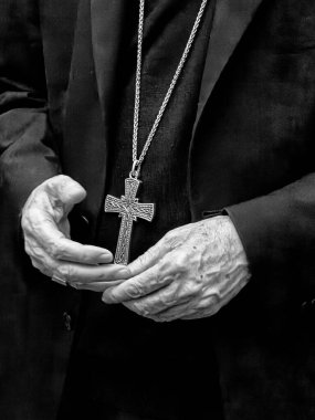 Siyah cübbeli bir kardinalin iki eli yüksek rütbeli rahiplerin inanç ve papalık otoritesinin sembolü olarak göğüs kafesini çerçeveler. Siyah ve beyaz..