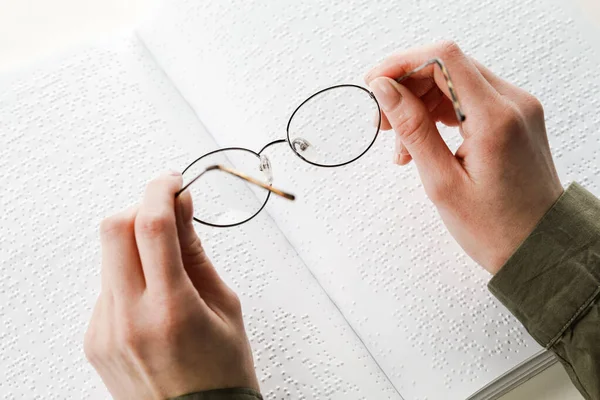 A woman holds black-rimmed glasses against the background of an open textbook reading books in Braille.
