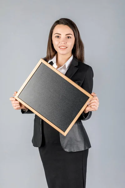 Young business woman in dark suite with black board in frame on grey background. Office concept