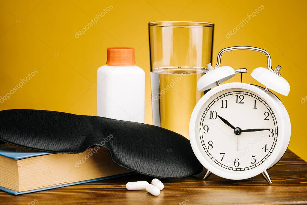 A mask, a glass of water, a book, pills, and an alarm clock. Restoring sleep patterns and treating insomnia.