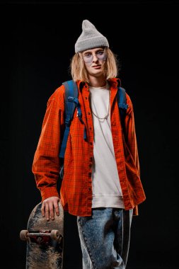 Stüdyoda kaykay tutan şık bir adamın portresi. Siyah arka planda poz veren gülümseyen bir kaykaycının yakın çekimi.