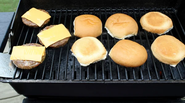 Hamburger patties with processed cheese slices cooking on an outdoor table top barbecue.  Buns toasting alongside — Stock Photo, Image