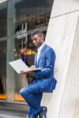 New York 'ta çalışan, mavi takım elbiseli, beyaz gömlekli, ofis binasının önünde köşesinde duran, dizüstü bilgisayarını okuyan genç bir Afro-Amerikan iş adamı.