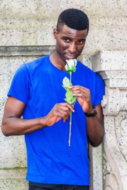 Genç Afrikalı Amerikalı adam New York 'ta seni özlüyor, mavi kısa kollu tişört giyiyor, kampüsün klasik duvarının köşesinde duruyor, elinde beyaz bir gül çiçeği tutuyor, başını eğip, çiçekleri kokluyor.