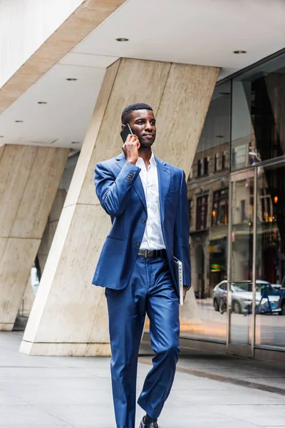 Young African American businessman traveling, working in New York City, wearing blue suit, white shirt, walking on street outside office building with glass windows, talking on cell phone