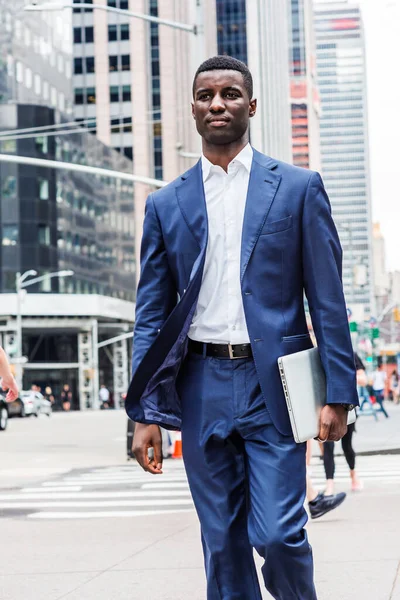 Young African American Businessman traveling in New York City, wearing blue suit, white undershirt, carrying laptop computer, walking on busy street with high buildings in Midtown of Manhattan