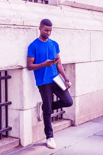 Young African American college student studying in New York City, wearing blue T suit, black pants, sneakers, holding laptop computer, standing against wall outside office building, texting on phone
