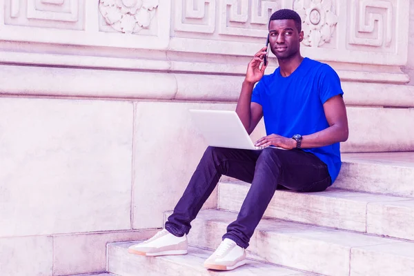 Young African American College Student studying in New York, wearing blue T shirt, black pants, sneakers, wristwatch, sitting on stairs on campus, working on laptop computer, talking on cell phone