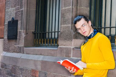 Genç İspanyol asıllı Amerikalı adam New York 'ta kırmızı kitap okuyor, saçı topuz gibi, gözlüklü, sarı uzun kollu tişörtlü, boynunda küçük siyah atkısı olan, duvarın yanında duran, düşünen, düşünen.