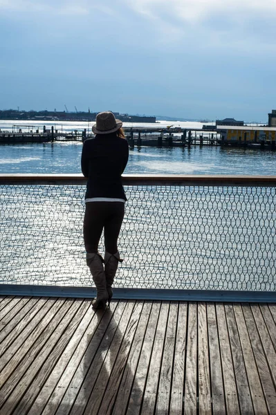 In the sunshine, a woman dressing in black and wearing a hat is standing on the dock, watching a beautiful scene.