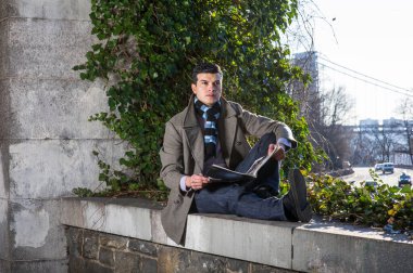 Genç bir adam büyük bir çerçevede oturuyor ve vahşi yeşil bitkilerin önünde bir dergi okuyor ve düşünüyor. Arka planda yolda giden arabalar ve büyük bir köprü var.