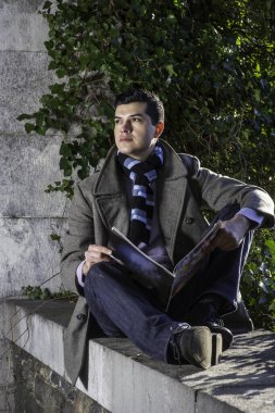 Genç bir adam büyük bir çerçevede oturuyor ve vahşi yeşil bitkilerin önünde bir dergi okuyor ve düşünüyor..