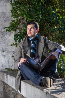 Genç bir adam büyük bir çerçevede oturuyor ve vahşi yeşil bitkilerin önünde bir dergi okuyor ve düşünüyor..