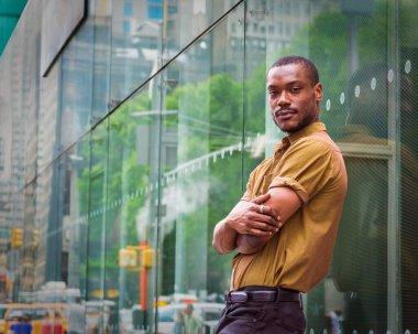 Siyah sakallı, kısa kollu yeşil gömlekli, siyah pantolonlu, kolları önde çapraz duran genç bir adam Manhattan, New York 'ta bir sokakta yansımalarıyla cam duvara dayanıyor.