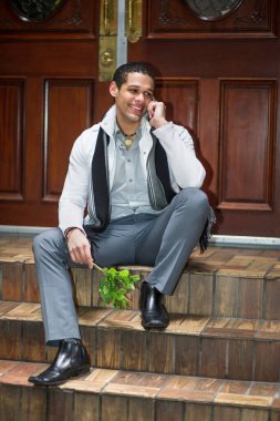 Elinde küçük yeşil bitkiler olan ve telefonla konuşan genç bir adam kapının önünde oturmuş seni bekliyor.